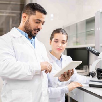 Confident experienced middle-eastern scientist in lab coat showing data on tablet to colleague while they examining test solution in laboratory