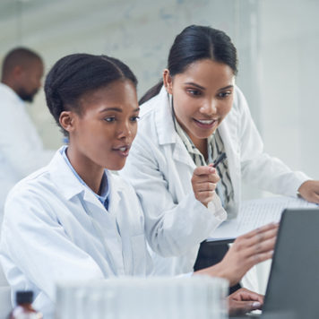 two women wearing white lab coats review results on computer